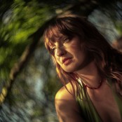 A close up image of a woman with wavy brown hair wearing a red necklace and green top
